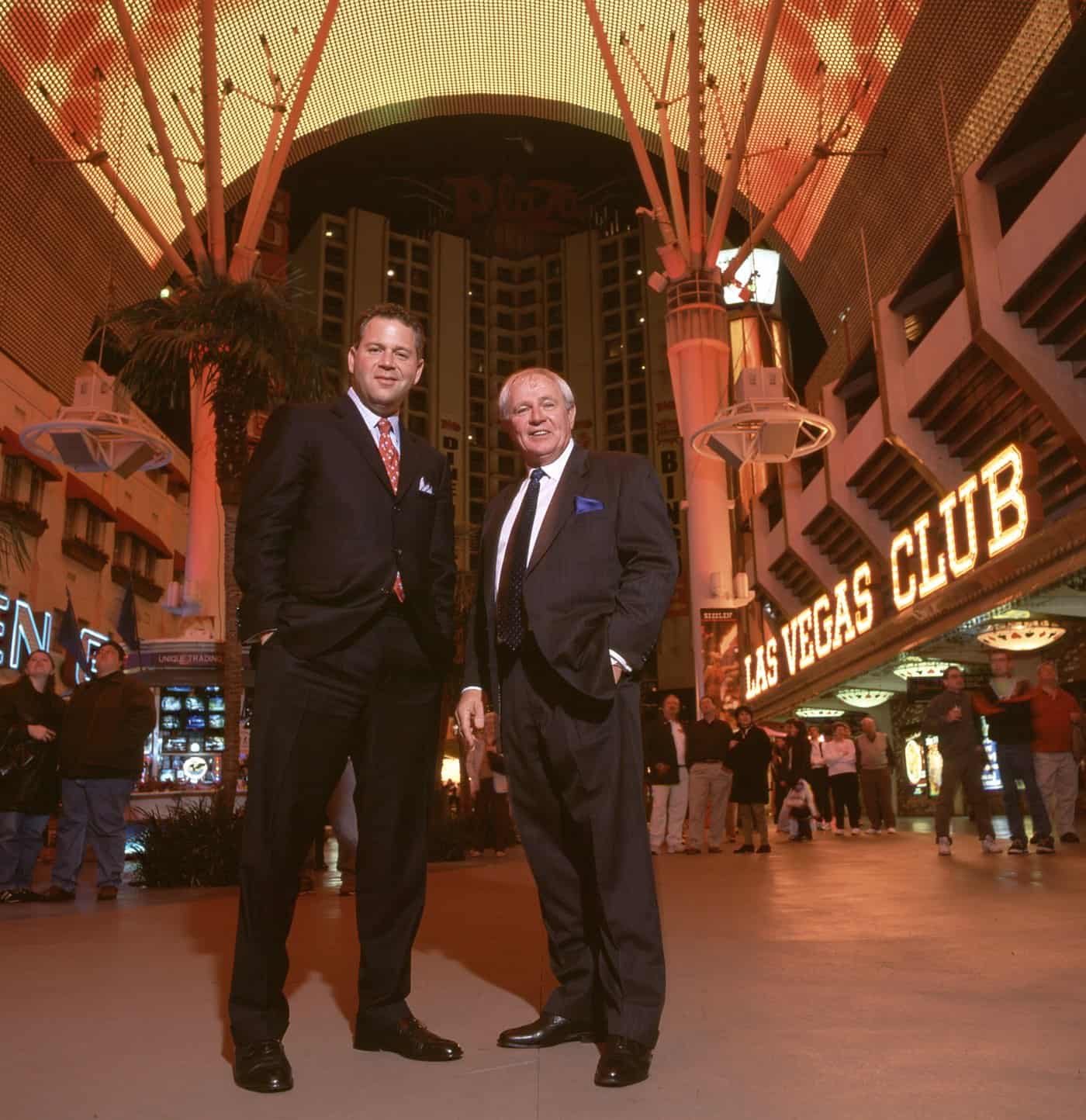 Dave Barrick and Stephen Crystal in dark suits, standing together beneath the lit Fremont Street Experience canopy with the Plaza tower rising behind them.