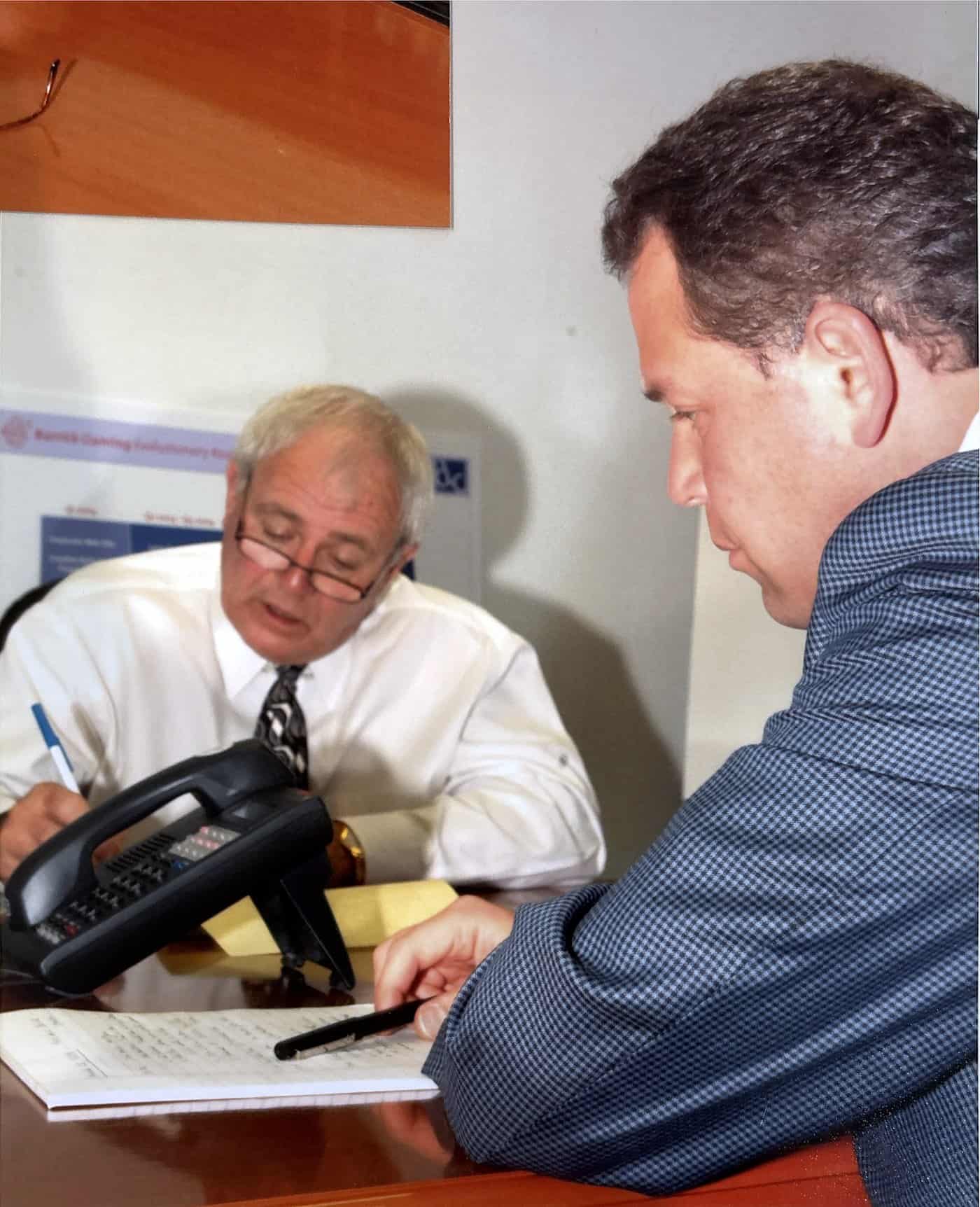 Two men at a desk, one signing paperwork while the other looks on.