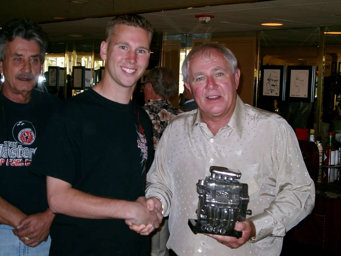 Two men shaking hands over a presented antique slot machine at an indoor gathering.